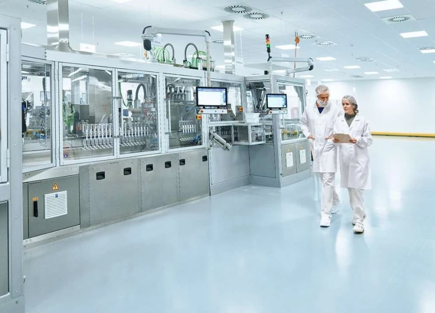 Two healthcare workers in lab coats walk past pharmaceutical manufacturing equipment inside a bright, sterile cleanroom environment.