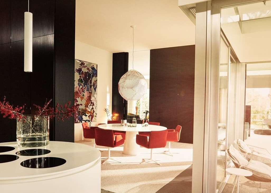 Interior view of a modern dining room featuring a white round table with red chairs, complemented by abstract art and globe lighting.