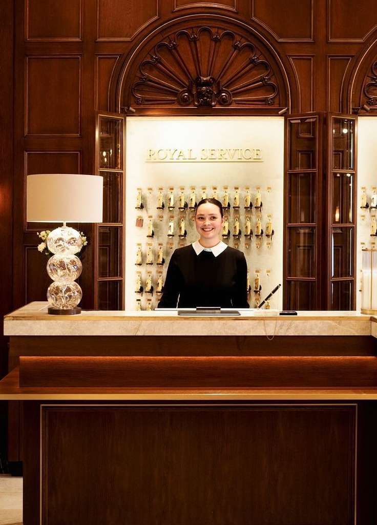 Hotel front desk scene featuring a smiling woman in a black dress standing behind a wooden counter with a crystal lamp and key rack.