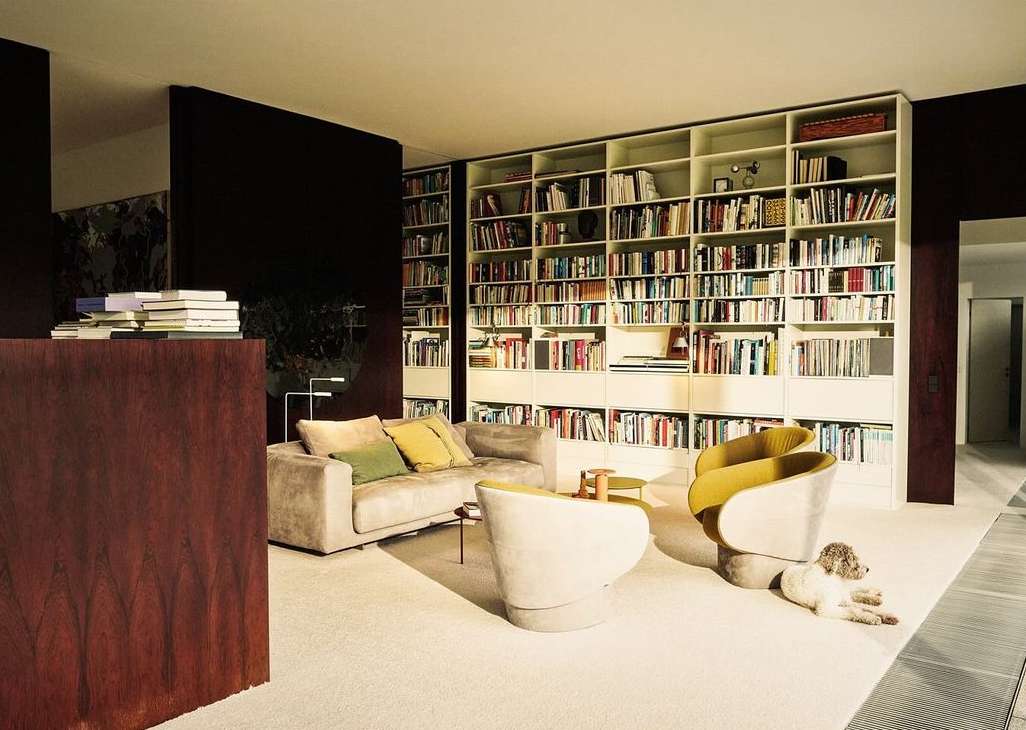 Interior living room view featuring a tan sofa with throw pillows, mid-century chairs, and a large built-in bookshelf filled with books.