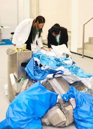 Two women in suits inspecting a pile of discarded trash bags and debris in an indoor setting, with a metal waste bin nearby.