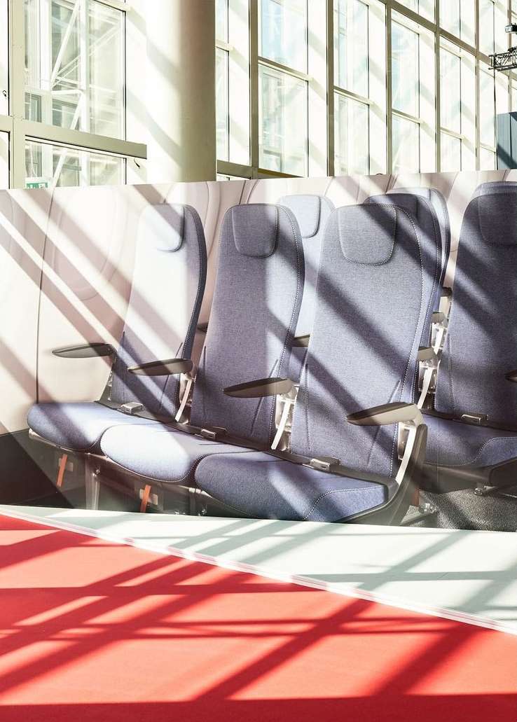 Interior view of rows of airplane seats with overhead sunlight casting striped shadows across the seats and red flooring in the foreground.