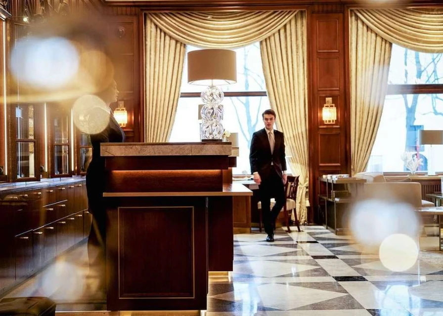 Hotel lobby scene featuring a man in a suit walking, with a woman standing at the reception desk and elegant interior design.