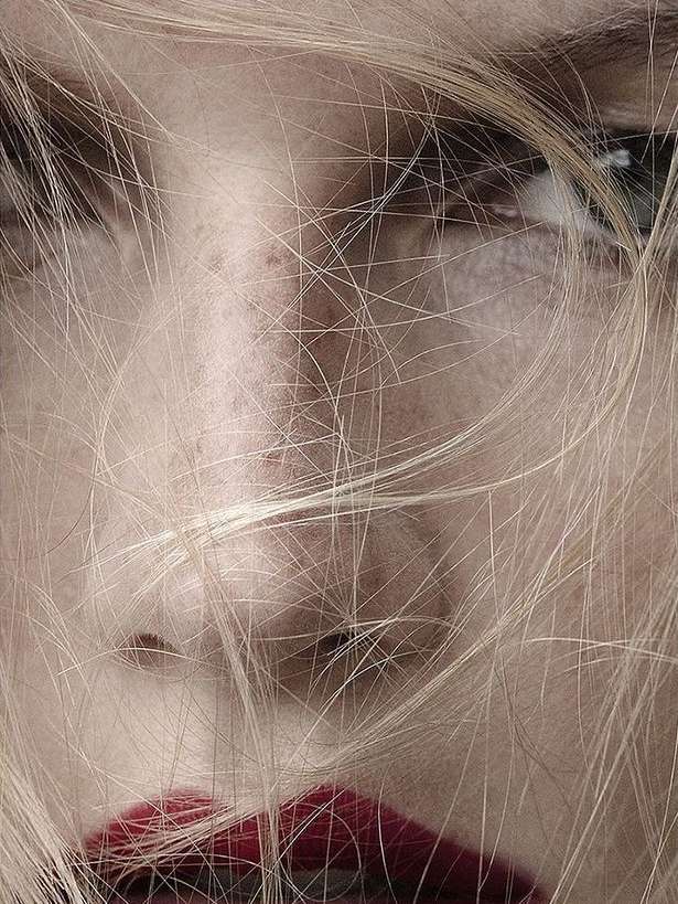 Close-up portrait of a blonde woman's face with strands of hair partially obscuring her face, with visible eyes, nose, and red lips.
