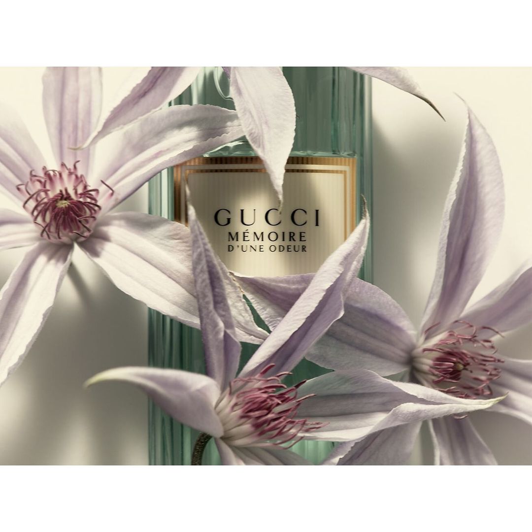 Close-up of a 'Gucci Mémoire d'une Odeur' perfume bottle surrounded by delicate purple and white clematis flowers on a light surface.