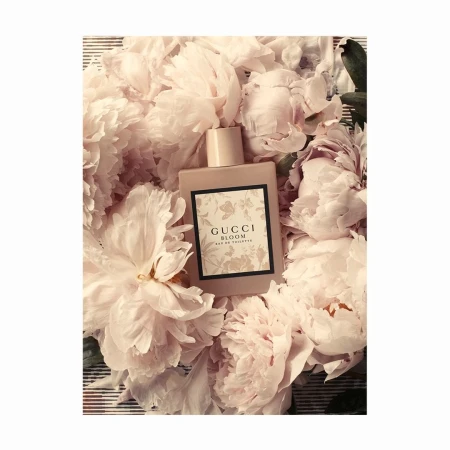 Gucci Bloom Eau de Toilette perfume bottle nestled in a bed of light pink peony flowers, beauty product shot in a close-up setting.