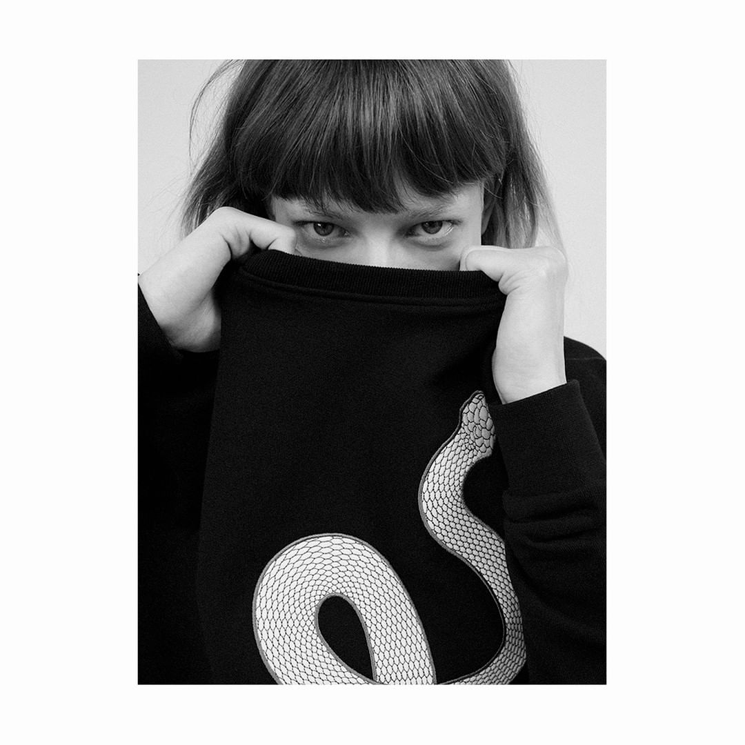Monochrome portrait of a woman partially covering her face with a black sweater featuring a coiled snake design, set against a plain backdrop.