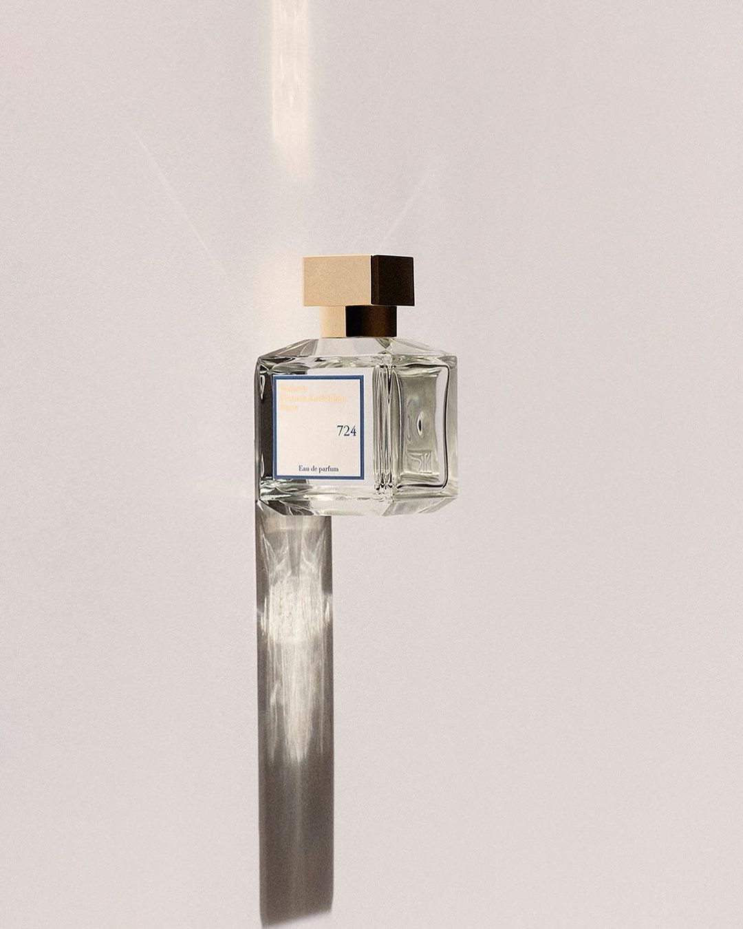 A clear glass perfume bottle with a square gold cap positioned vertically against a minimalist white surface with a long shadow.