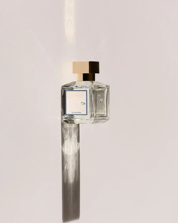 A clear glass perfume bottle with a square gold cap positioned vertically against a minimalist white surface with a long shadow.