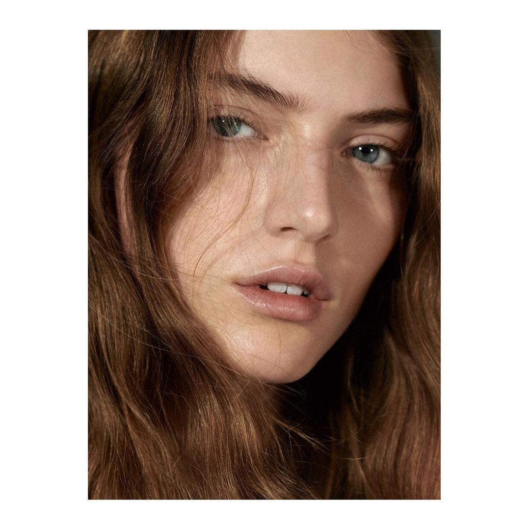 Close-up beauty shot of a woman with light skin, natural makeup, and wavy brown hair, captured in soft, diffused lighting indoors.