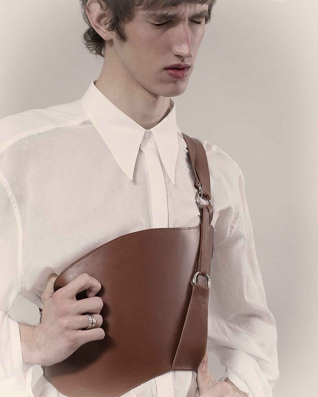 Young man in a white collared shirt wearing a brown leather asymmetrical chest harness by Berengere Valognes