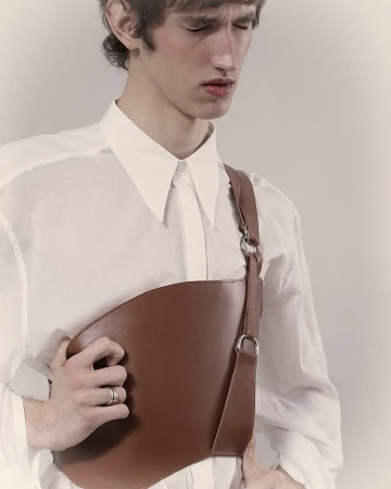 Young man in a white collared shirt wearing a brown leather asymmetrical chest harness by Berengere Valognes