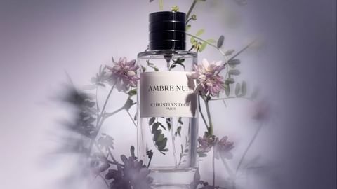A clear glass bottle of Dior's Ambre Nuit perfume is positioned in front of a blurred background with delicate purple flowers and green leaves.