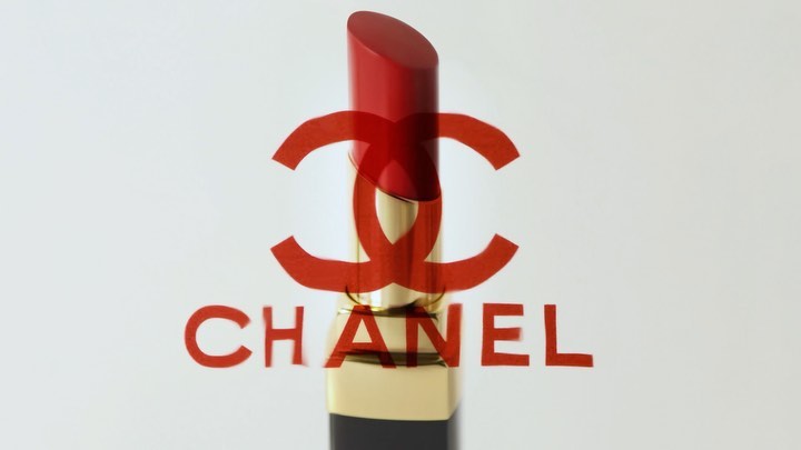 A red lipstick tube with a golden base sits in front of a translucent red Chanel logo on a bright white background in studio lighting.