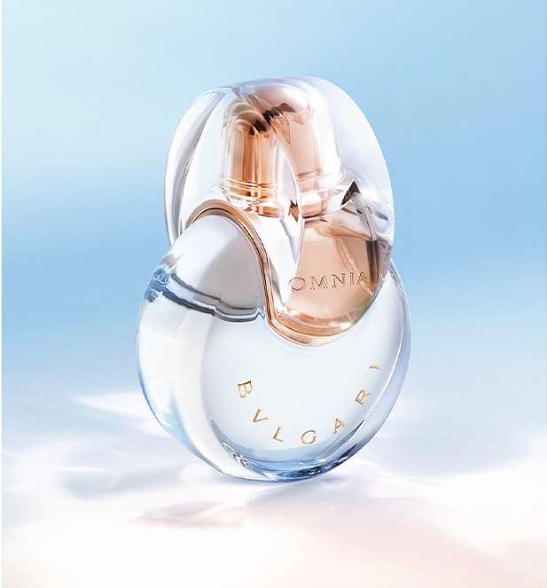 Clear glass Bulgari Omnia perfume bottle with gold accents on a light blue background with cloud-like formations, in a studio shot.