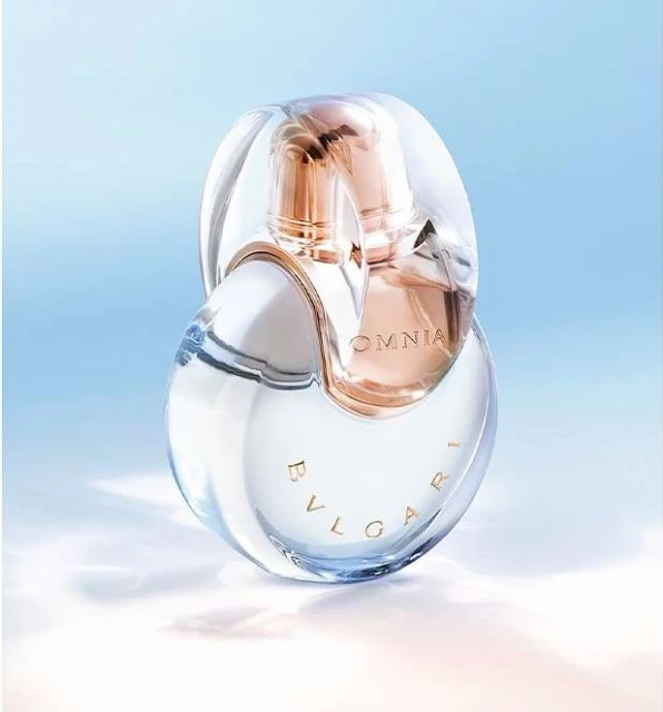 Clear glass Bulgari Omnia perfume bottle with gold accents on a light blue background with cloud-like formations, in a studio shot.