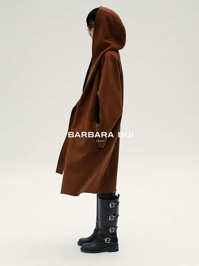 Fashion shot featuring a woman in a brown hooded coat and black buckle boots against a bright studio backdrop, showcasing style and design.