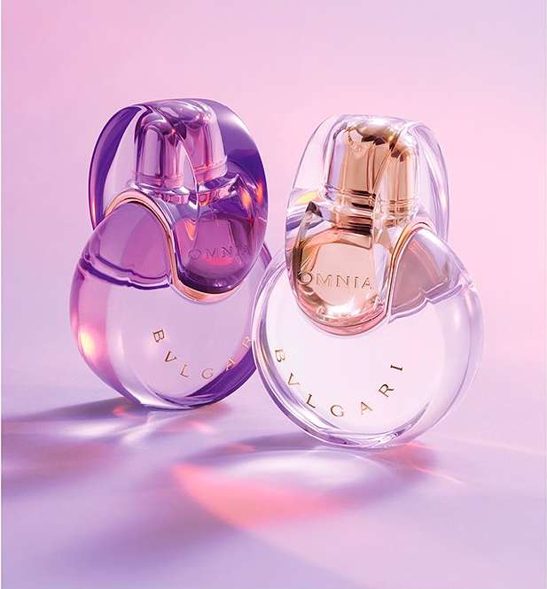 Two Bulgari Omnia perfume bottles, one purple and one gold, sit on a purple surface with a soft pink and purple gradient background. 