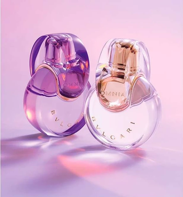 Two Bulgari Omnia perfume bottles, one purple and one gold, sit on a purple surface with a soft pink and purple gradient background. 