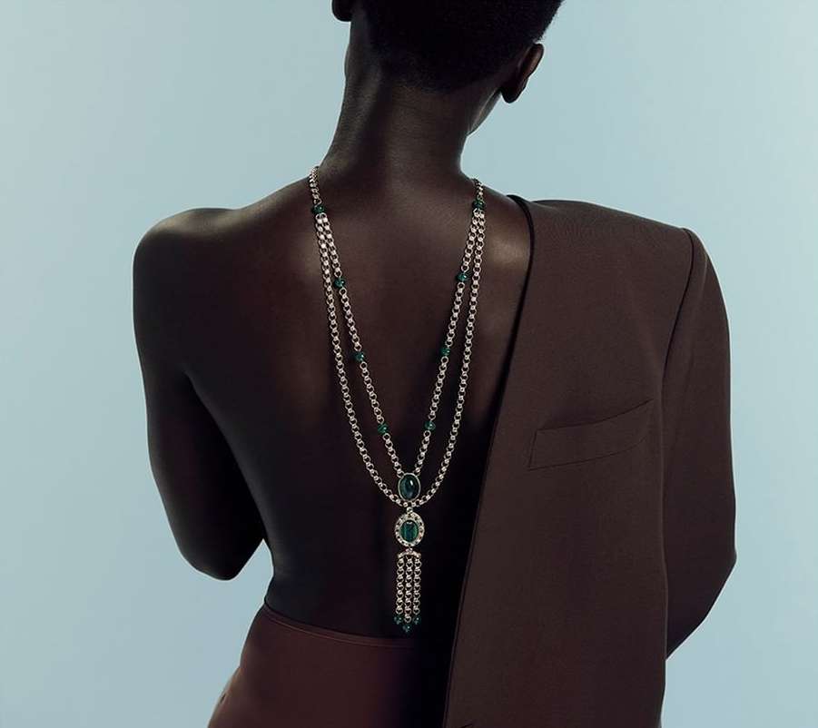 Elegant back view of a dark-skinned adult female adorned with a silver and emerald necklace, complemented by a brown blazer and skirt.
