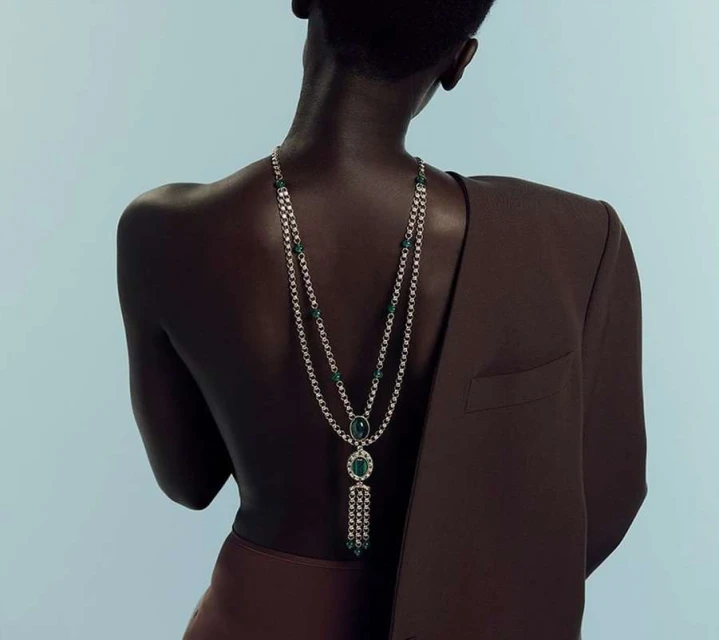 Elegant back view of a dark-skinned adult female adorned with a silver and emerald necklace, complemented by a brown blazer and skirt.