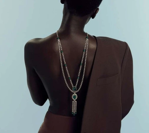 Elegant back view of a dark-skinned adult female adorned with a silver and emerald necklace, complemented by a brown blazer and skirt.