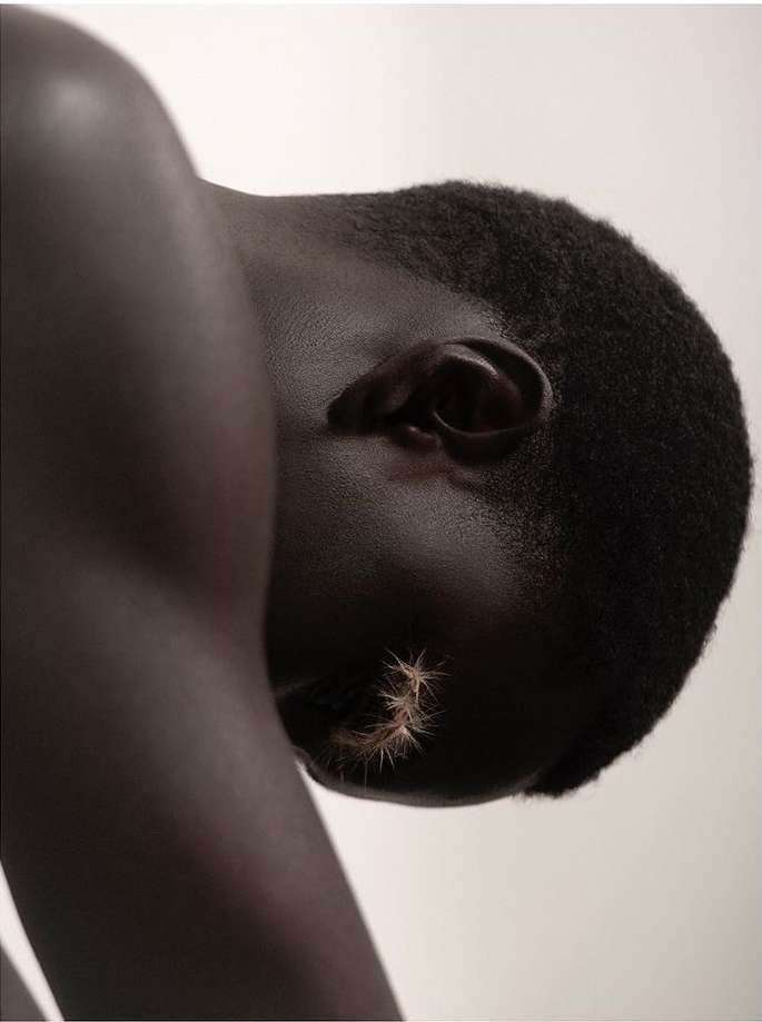 Close-up portrait of a dark-skinned adult with short hair and spiky decorative elements applied to the cheek against a plain background.