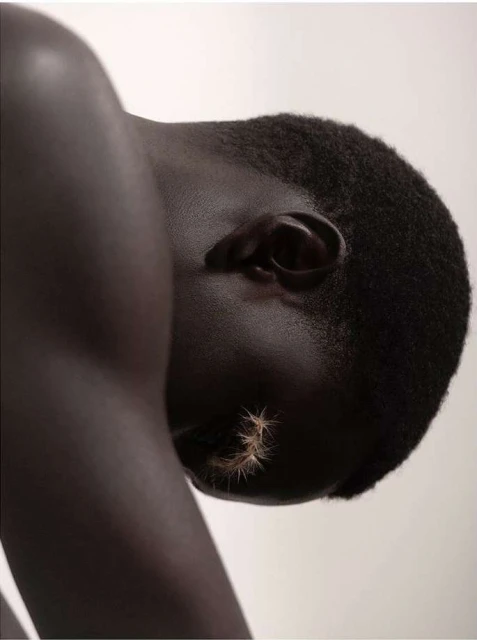 Close-up portrait of a dark-skinned adult with short hair and spiky decorative elements applied to the cheek against a plain background.