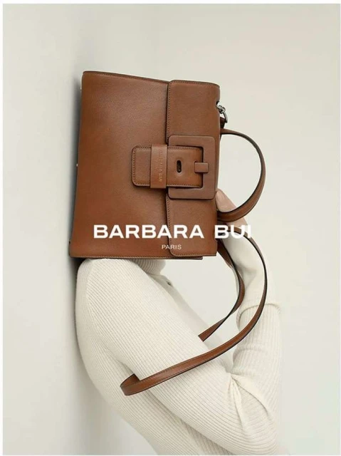 Brown leather Barbara Bui handbag with buckle detail, held by a person wearing a cream turtleneck sweater against a light background.