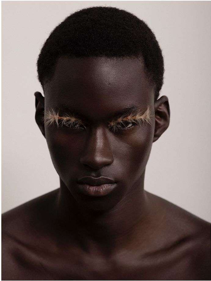 Close-up portrait of a black male model with dramatic light-brown false eyelashes against a plain background, beauty and fashion.