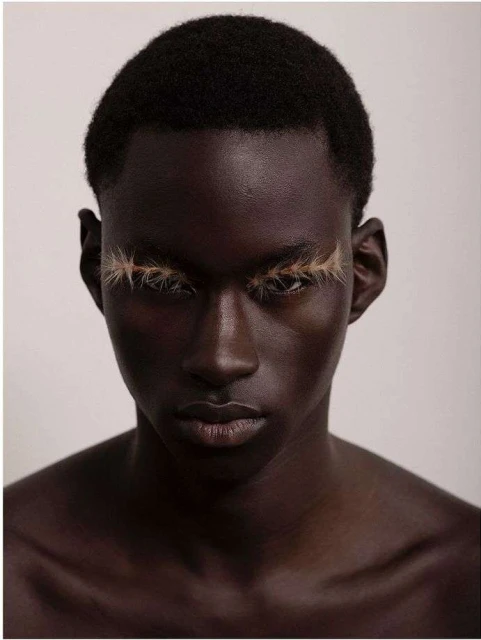 Close-up portrait of a black male model with dramatic light-brown false eyelashes against a plain background, beauty and fashion.