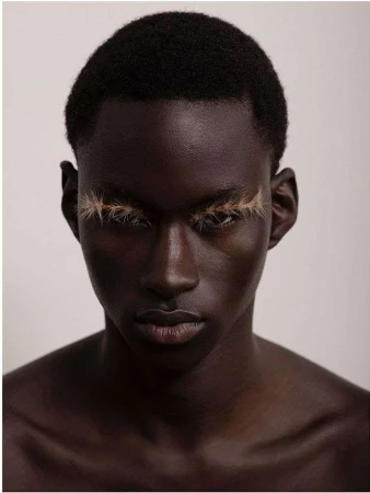 Close-up portrait of a black male model with dramatic light-brown false eyelashes against a plain background, beauty and fashion.