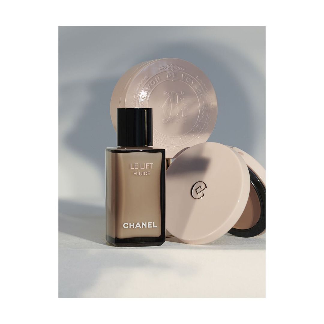 Still life of Chanel Le Lift Fluide skincare bottle, round soap, and open compact against a gradient light blue background.
