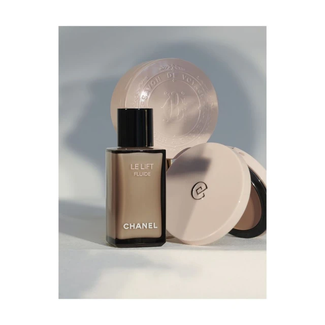 Still life of Chanel Le Lift Fluide skincare bottle, round soap, and open compact against a gradient light blue background.