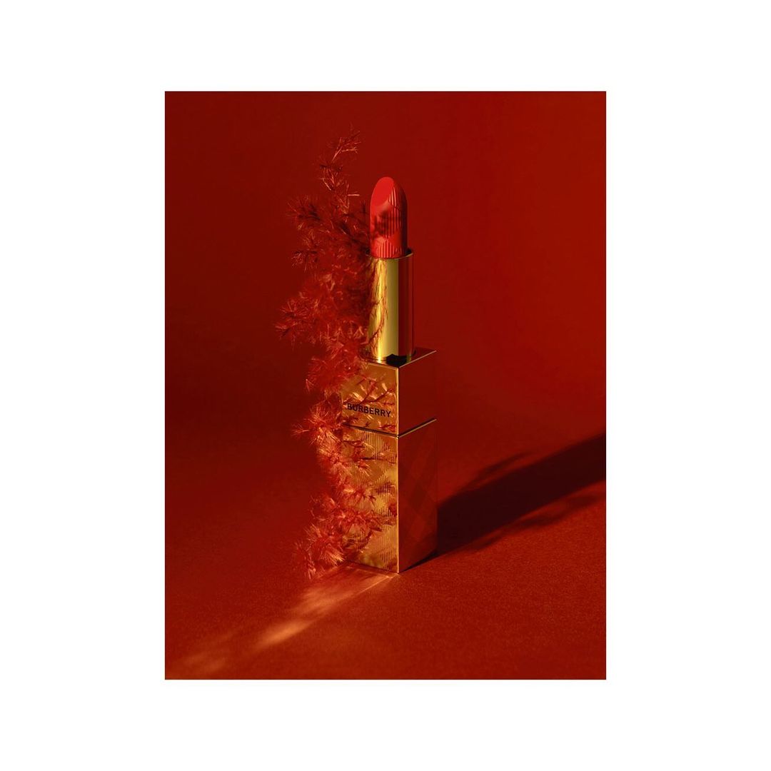 Close-up studio shot of gold lipstick tube with red lipstick, accented by soft red foliage against a deep red background.