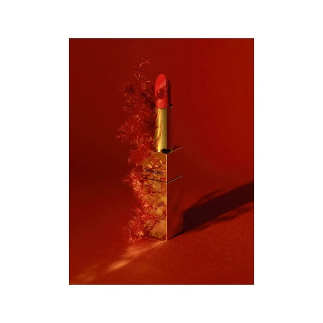Close-up studio shot of gold lipstick tube with red lipstick, accented by soft red foliage against a deep red background.