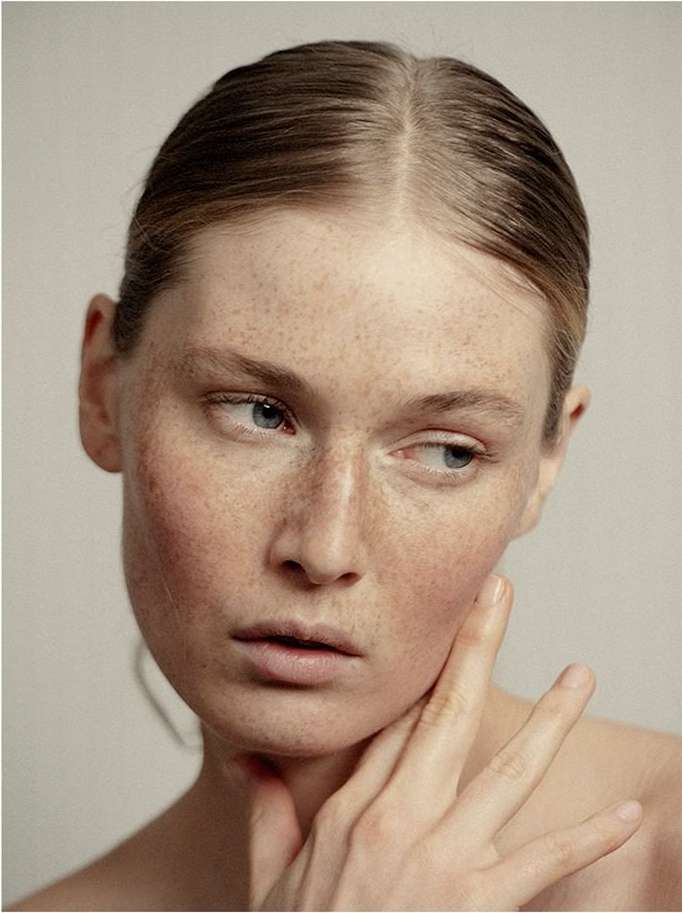 Close-up portrait of a fair-skinned young woman with freckles, hand near her face, gazing off to the side against a neutral background.