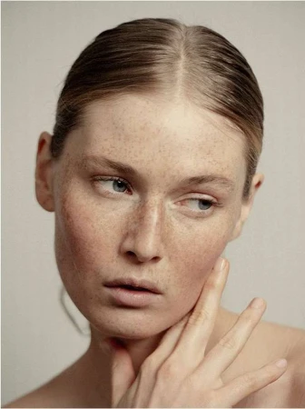 Close-up portrait of a fair-skinned young woman with freckles, hand near her face, gazing off to the side against a neutral background.