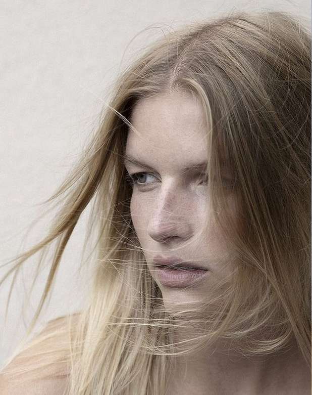 Close-up portrait of a blonde adult female with windblown hair against a neutral background in a beauty and fashion context.