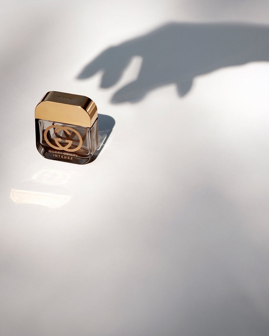 Gucci Guilty Intense perfume bottle with gold cap, illuminated by soft, natural light casting hand shadow on white background.