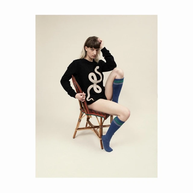 A person poses on a wicker chair, wearing a black sweater with a snake design and blue socks in a neutral studio setting.
