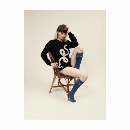 A person poses on a wicker chair, wearing a black sweater with a snake design and blue socks in a neutral studio setting.