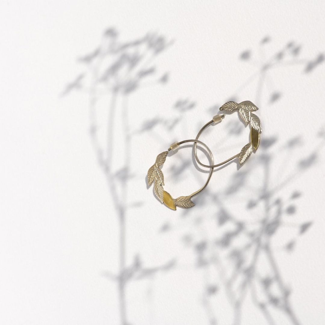 Pair of gold hoop earrings with leaf details on white background, casting soft floral shadows. Jewelry product shot.