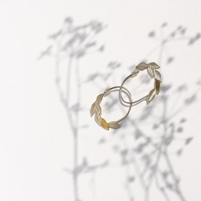 Pair of gold hoop earrings with leaf details on white background, casting soft floral shadows. Jewelry product shot.