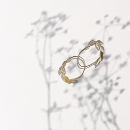 Pair of gold hoop earrings with leaf details on white background, casting soft floral shadows. Jewelry product shot.