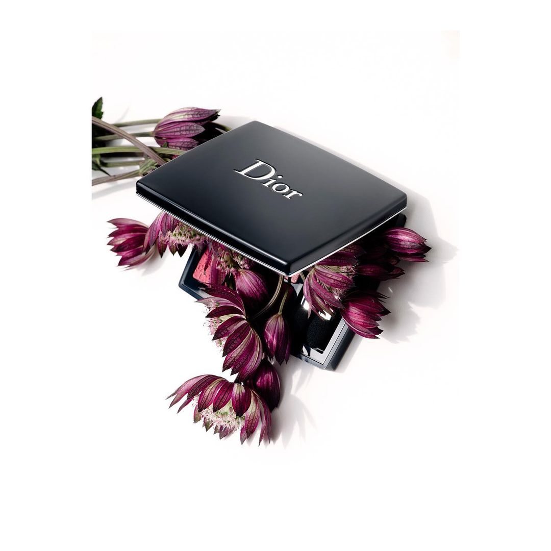 Dior makeup compact on white with flowers. Cosmetics product shot showcases makeup kit surrounded by purple flowers against a clean background.