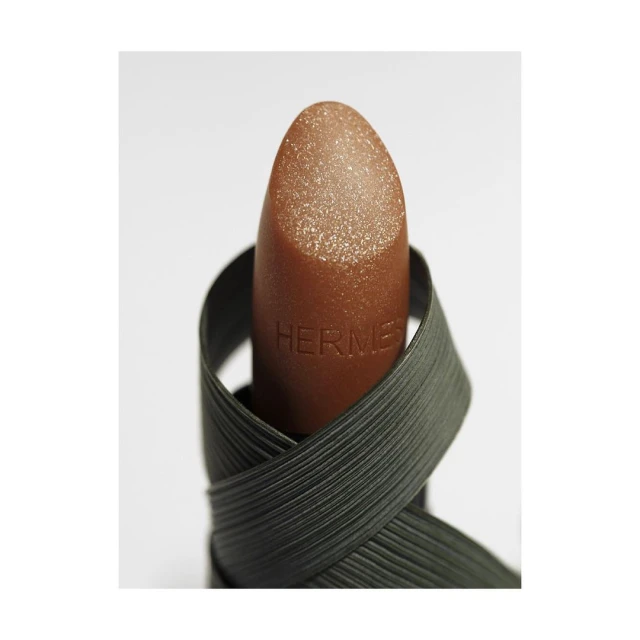 Close-up of bronze-colored Hermès lipstick wrapped with a dark green, textured ribbon against a bright white backdrop.