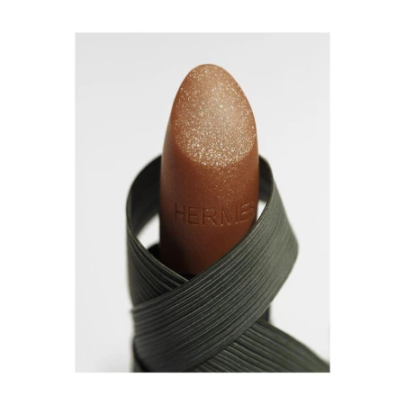 Close-up of bronze-colored Hermès lipstick wrapped with a dark green, textured ribbon against a bright white backdrop.