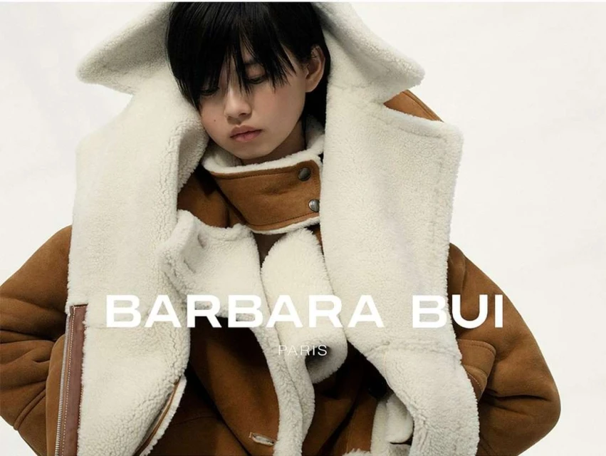 Woman poses in a Barbara Bui shearling coat against a bright background, showcasing high-fashion apparel with clear brand visibility.