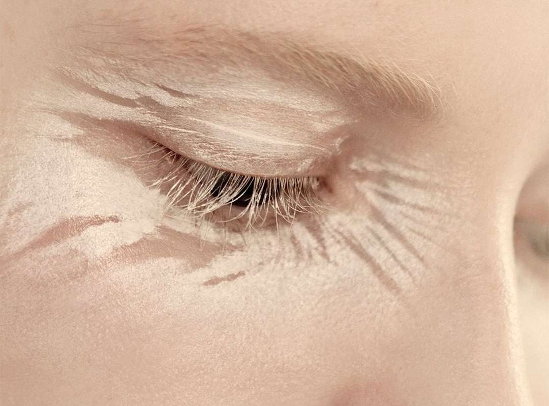 Close-up studio shot of a fair-skinned adult female's closed eye with long lashes, and artistic white paint designs on the face.